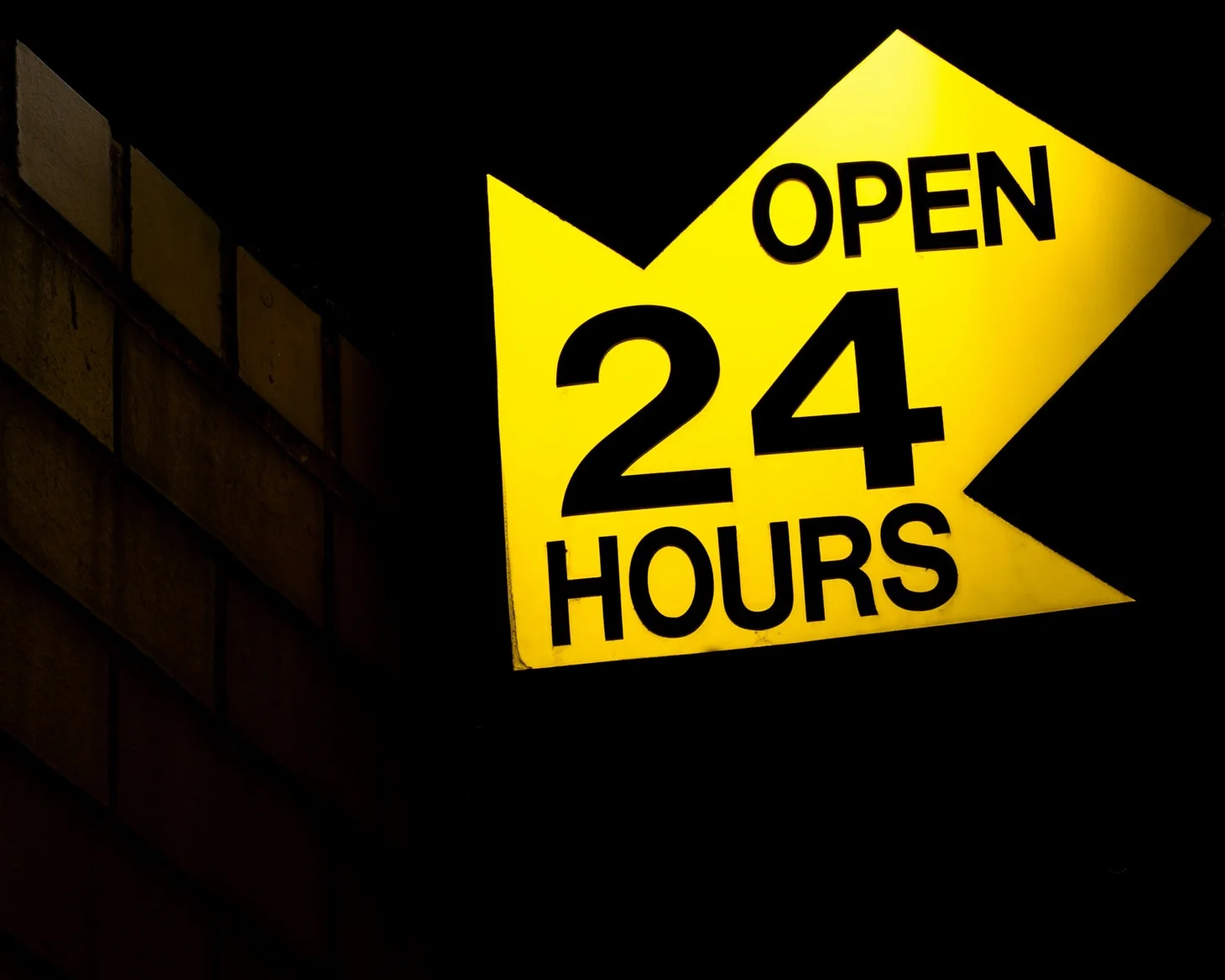 A yellow open 24 hours sign shining on the corner of a dark brick building at night.