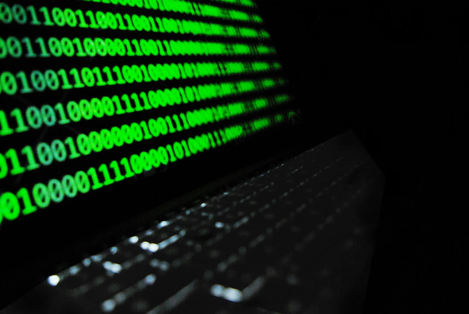 Green binary code glowing on a screen next to a laptop keyboard in the dark